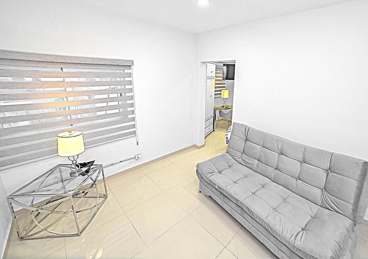 A living room with a grey sofa, a glass coffee table with a lamp, and a window with blinds.