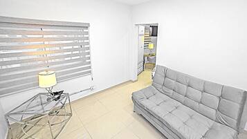 A living room with a grey sofa, a glass coffee table with a lamp, and a window with blinds.