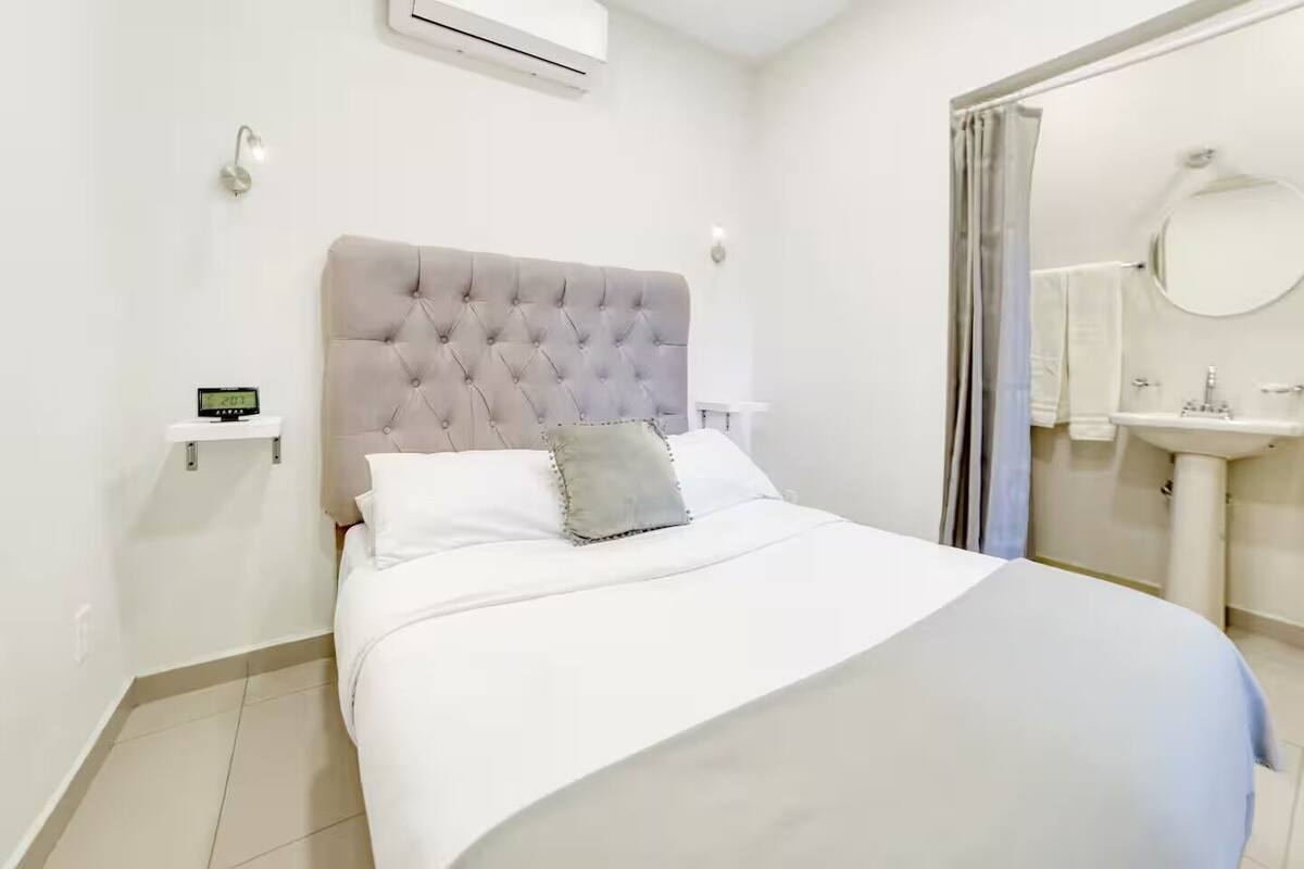A neatly made bed with a tufted headboard, a bedside clock, and a bathroom with a sink and mirror.