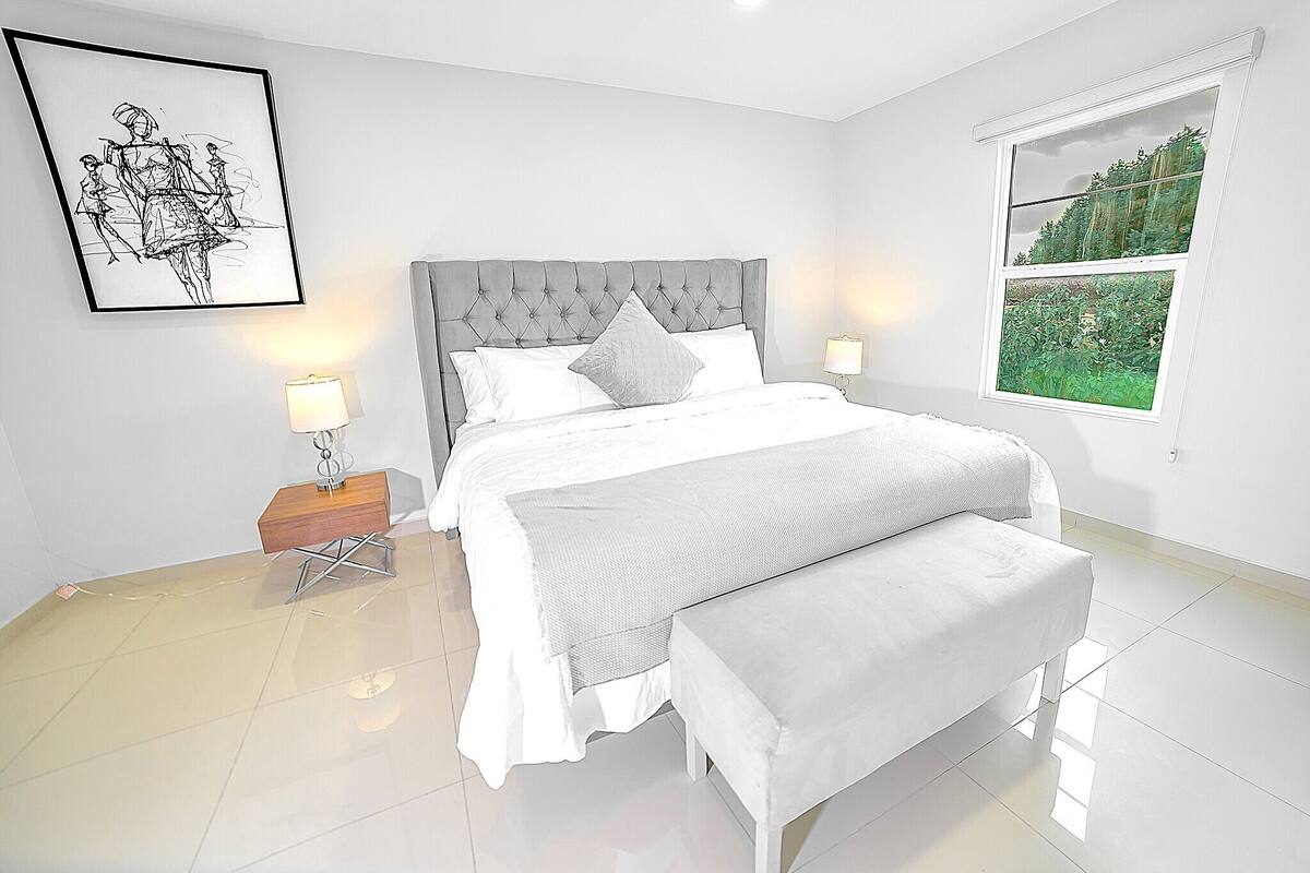 A bedroom with a large bed, a bedside table with a lamp, a bench, and a window with a view of greenery.