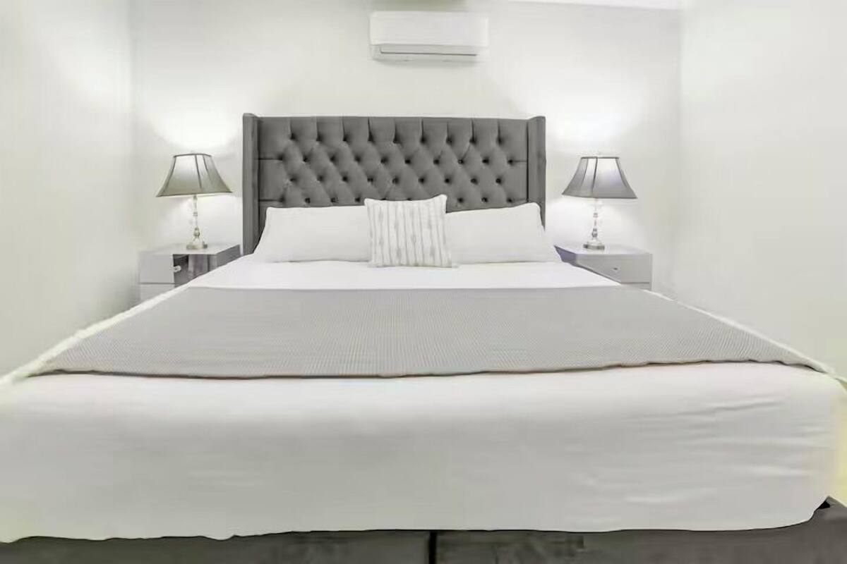 A neatly made bed with a tufted headboard, two bedside tables with lamps, and a wall-mounted air conditioner.