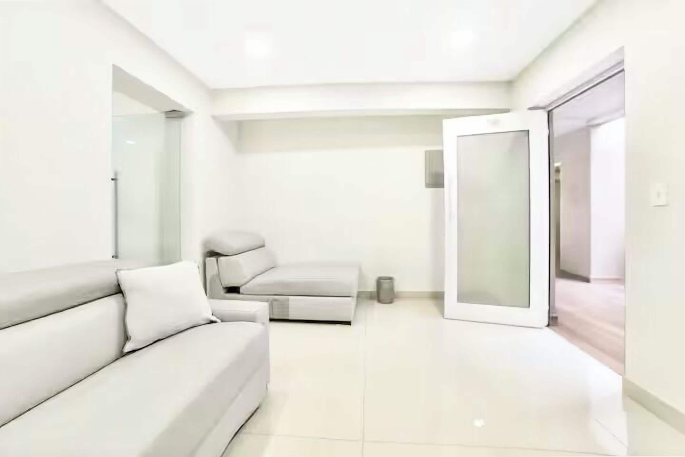 A modern, minimalist room with a white sofa, a glass door leading to another area, and a clean, tiled floor.