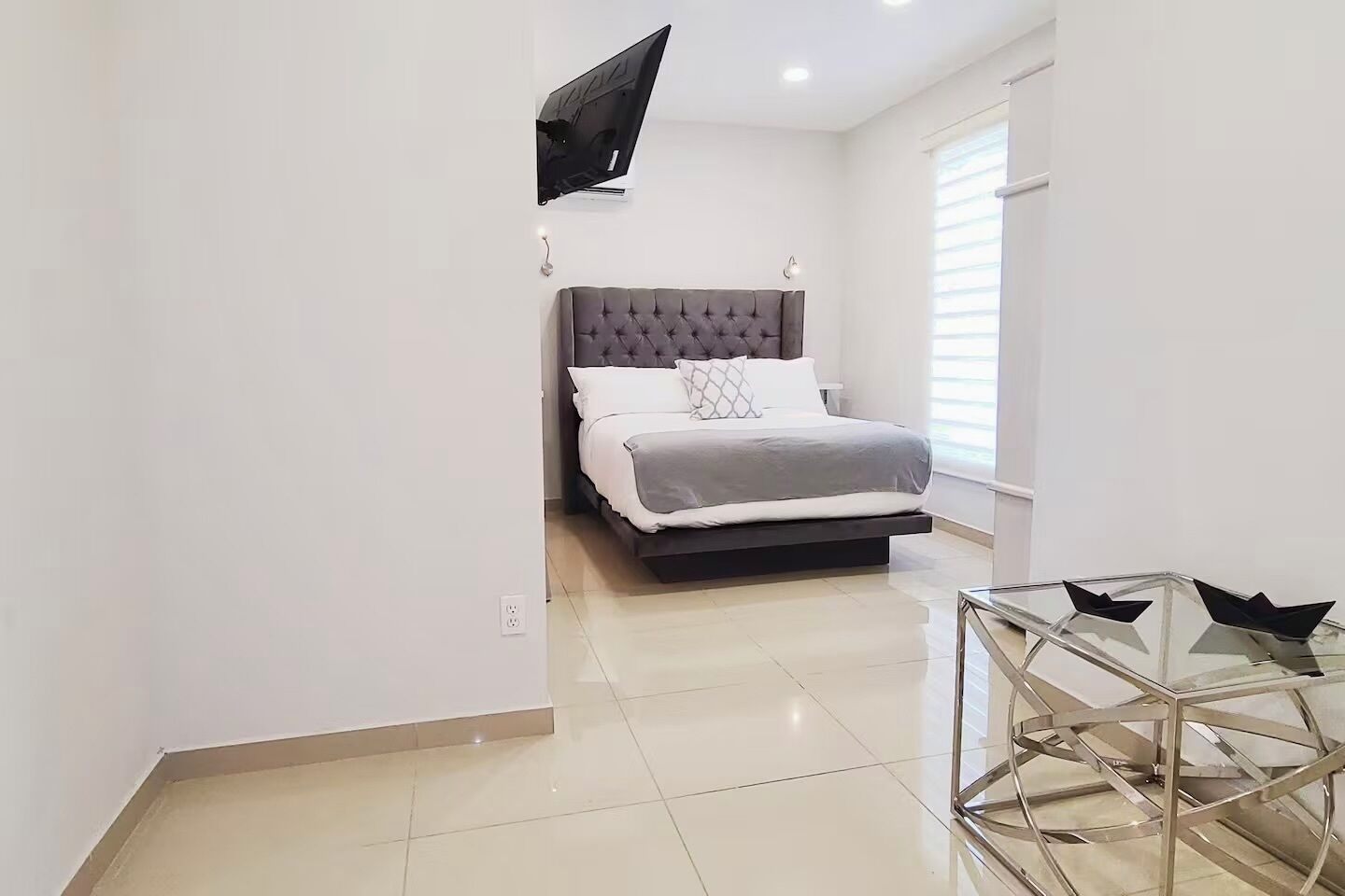 Un dormitorio moderno con cama, televisor montado en la pared y una mesa de vidrio con sillas.