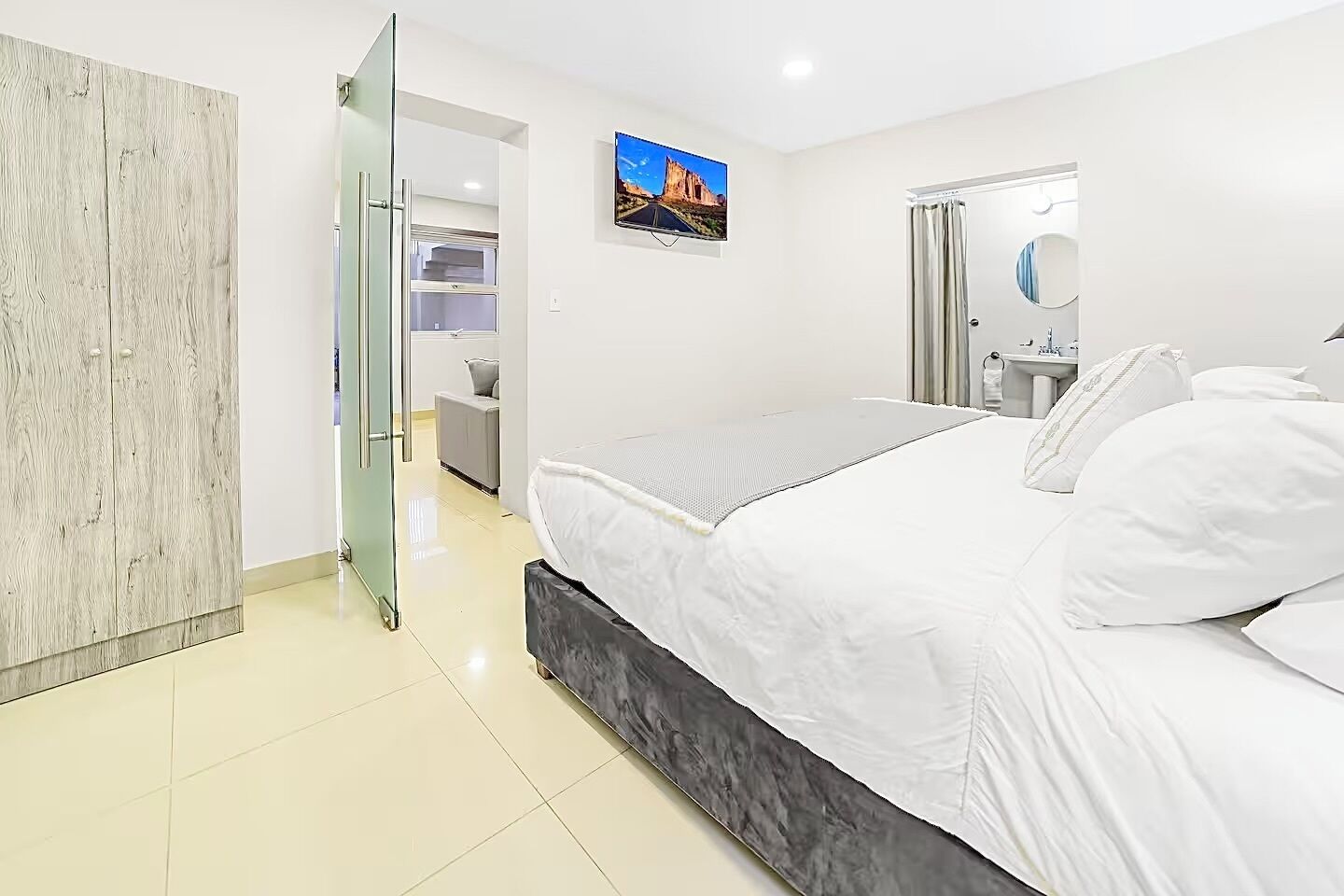 A modern bedroom with a bed, a wardrobe, a bathroom with a mirror, and a TV on the wall.
