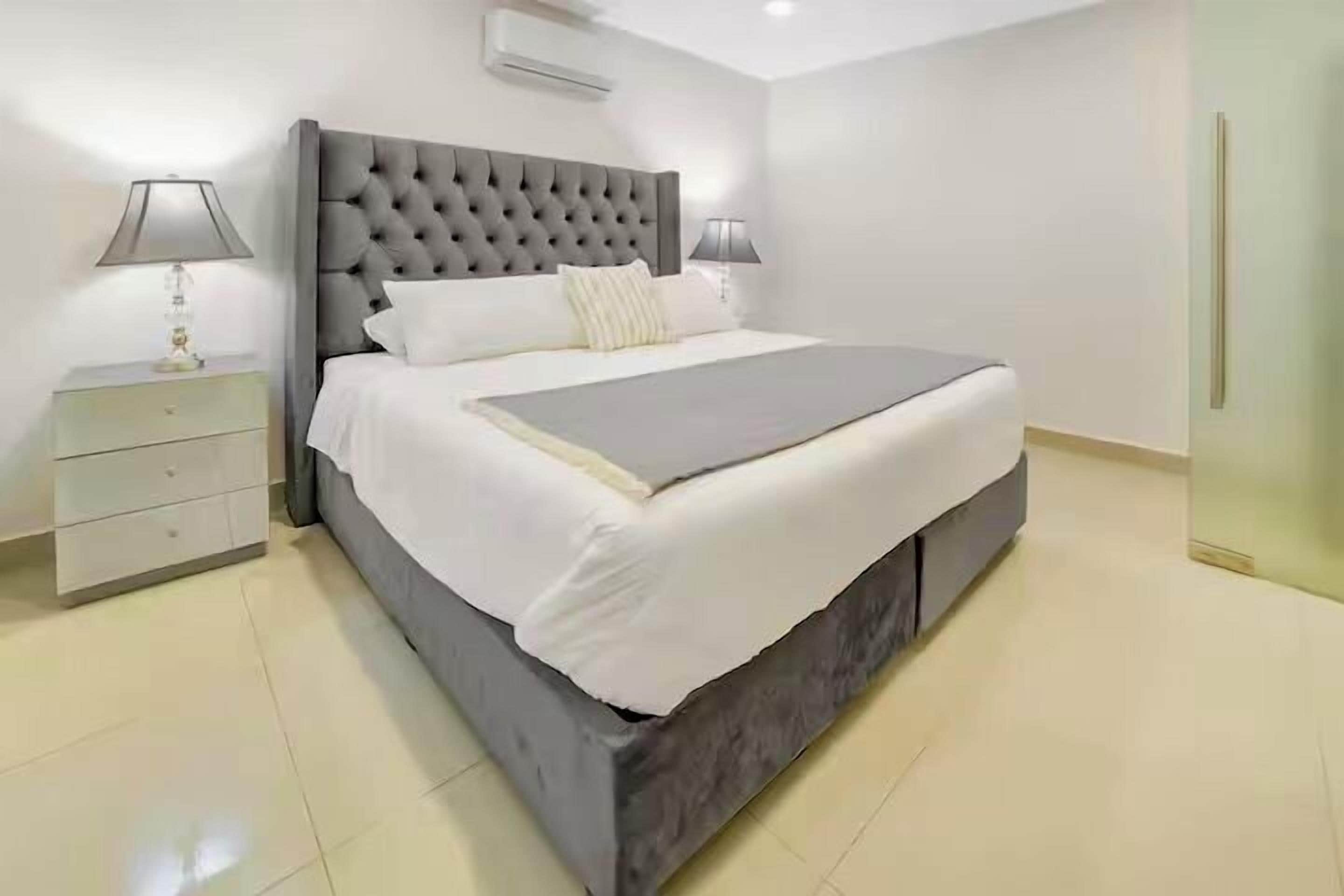 A modern bedroom with a tufted headboard, a large bed with white bedding, two bedside tables with lamps, and a small drawer unit.