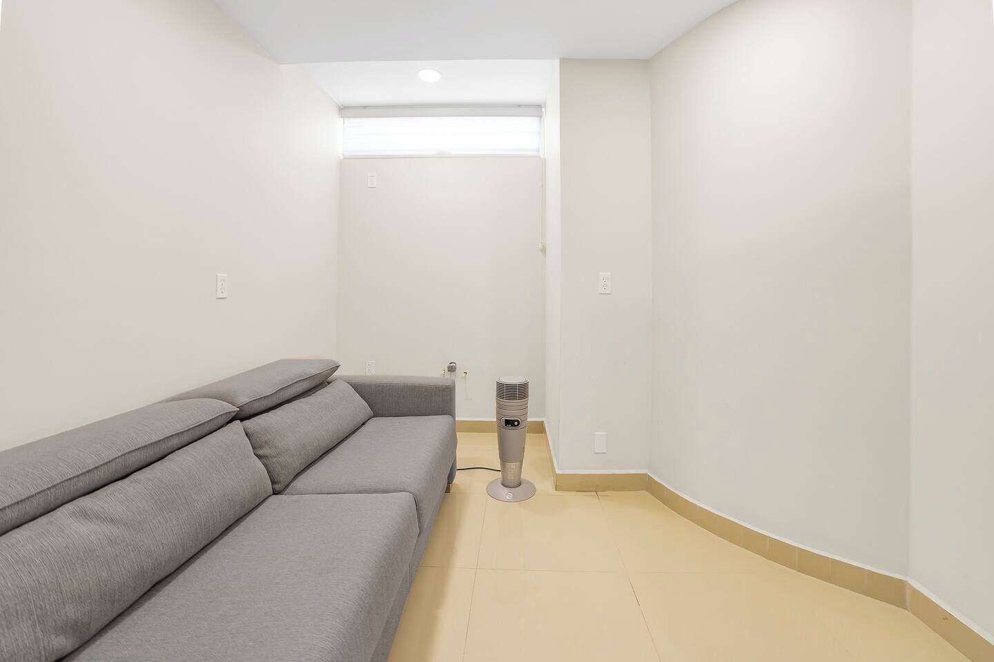 Une pièce avec un canapé gris, un radiateur et une fenêtre équipée de stores.