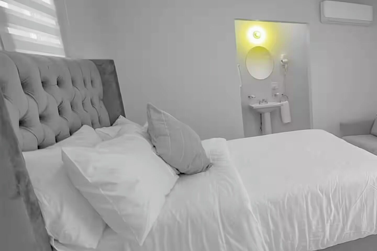 Quarto com cabeceira estofada, roupa de cama branca, mesa de cabeceira e espelho com luz na parede.