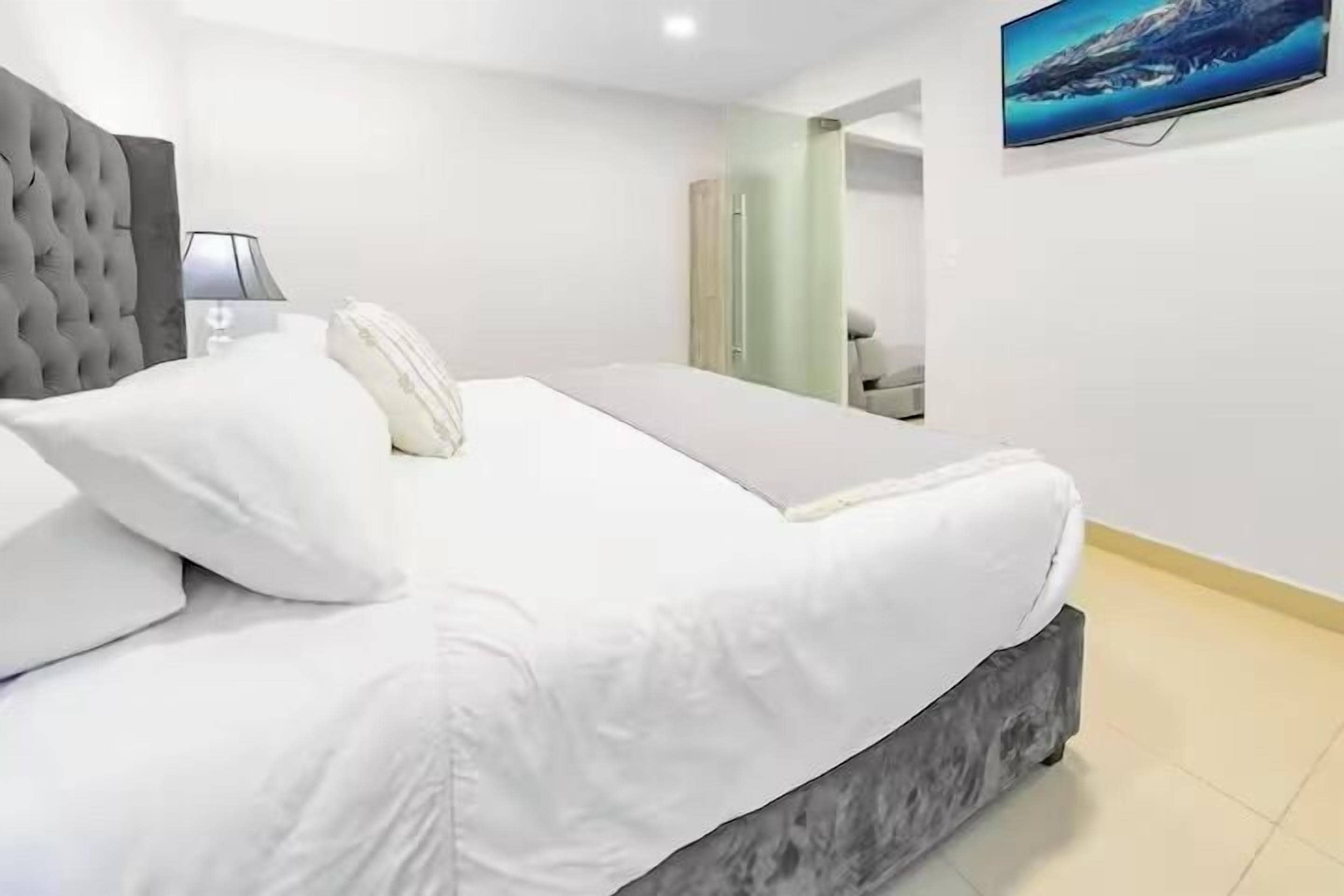 A modern bedroom with a large bed, a grey headboard, a bedside table with a lamp, and a painting on the wall.