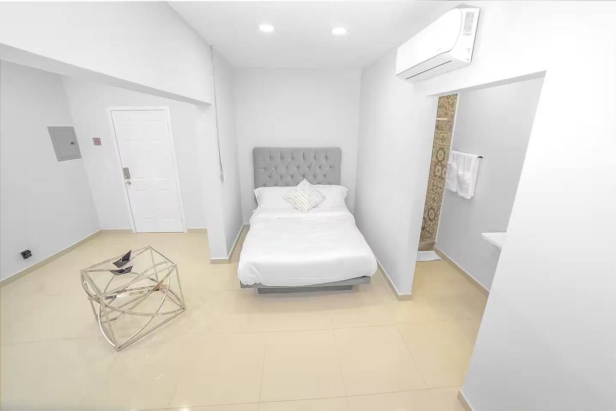 A modern bedroom with a bed, a wardrobe, an air conditioning unit, and a geometric glass table.