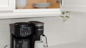 A kitchen with white cabinets, a coffee maker, and a white mug on a wooden board. - Cute, Cozy, and Curated South Hill Gem - 4 min to Manito Park (Spokane)
