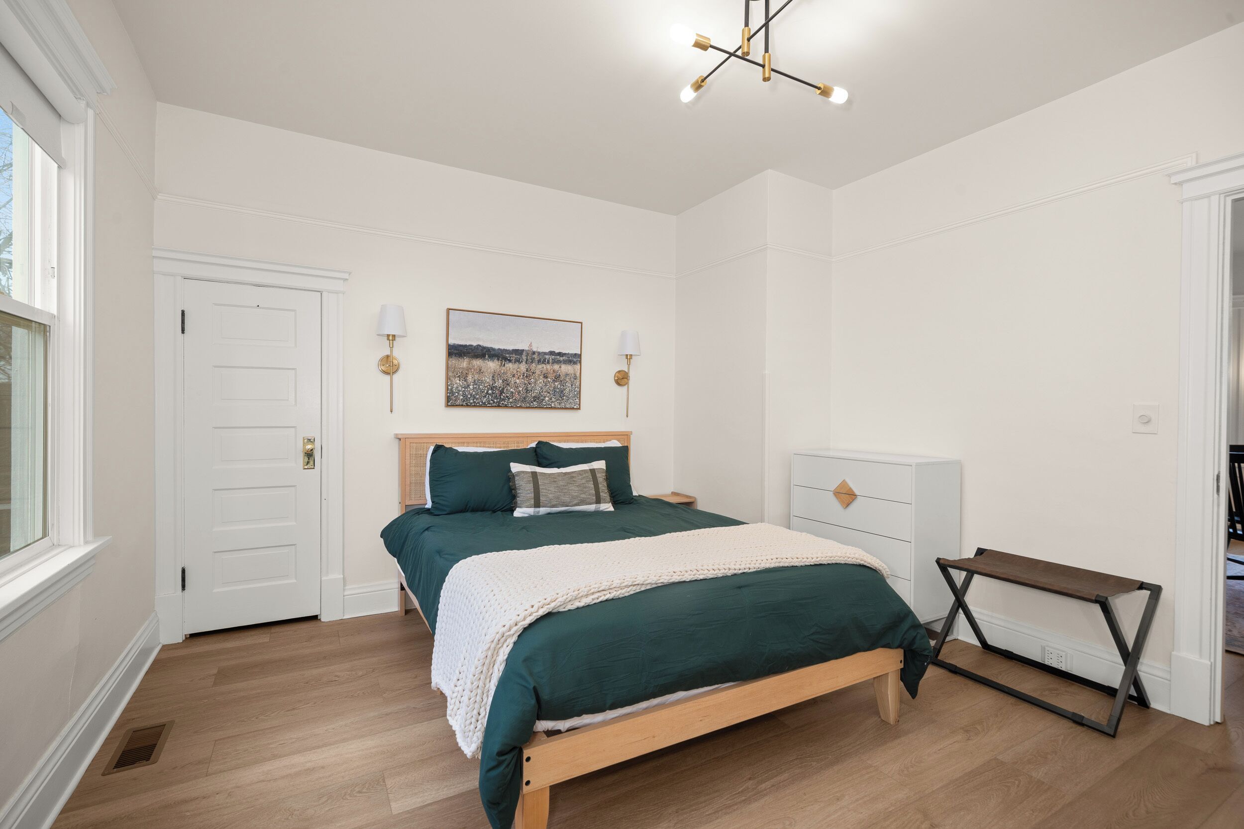Une chambre avec un lit en bois, des murs blancs, un luminaire au plafond et une fenêtre.