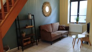Un salon avec un canapé marron, un escalier en bois, un miroir rond et une fenêtre avec des stores.