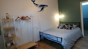 Une chambre avec un plafond en pente, un lit avec une literie à rayures, un luminaire suspendu et une étagère fixée au mur où sont posés des vêtements.