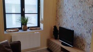 Un salon avec un canapé, un téléviseur posé sur un meuble en bois, une fenêtre avec des stores et un mur décoré d’un motif.