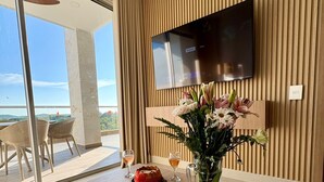 Een moderne woonkamer met een flatscreen televisie aan een houten wand, een houten salontafel met een bloevas, en uitzicht op het buitengebied door grote schuifdeuren.