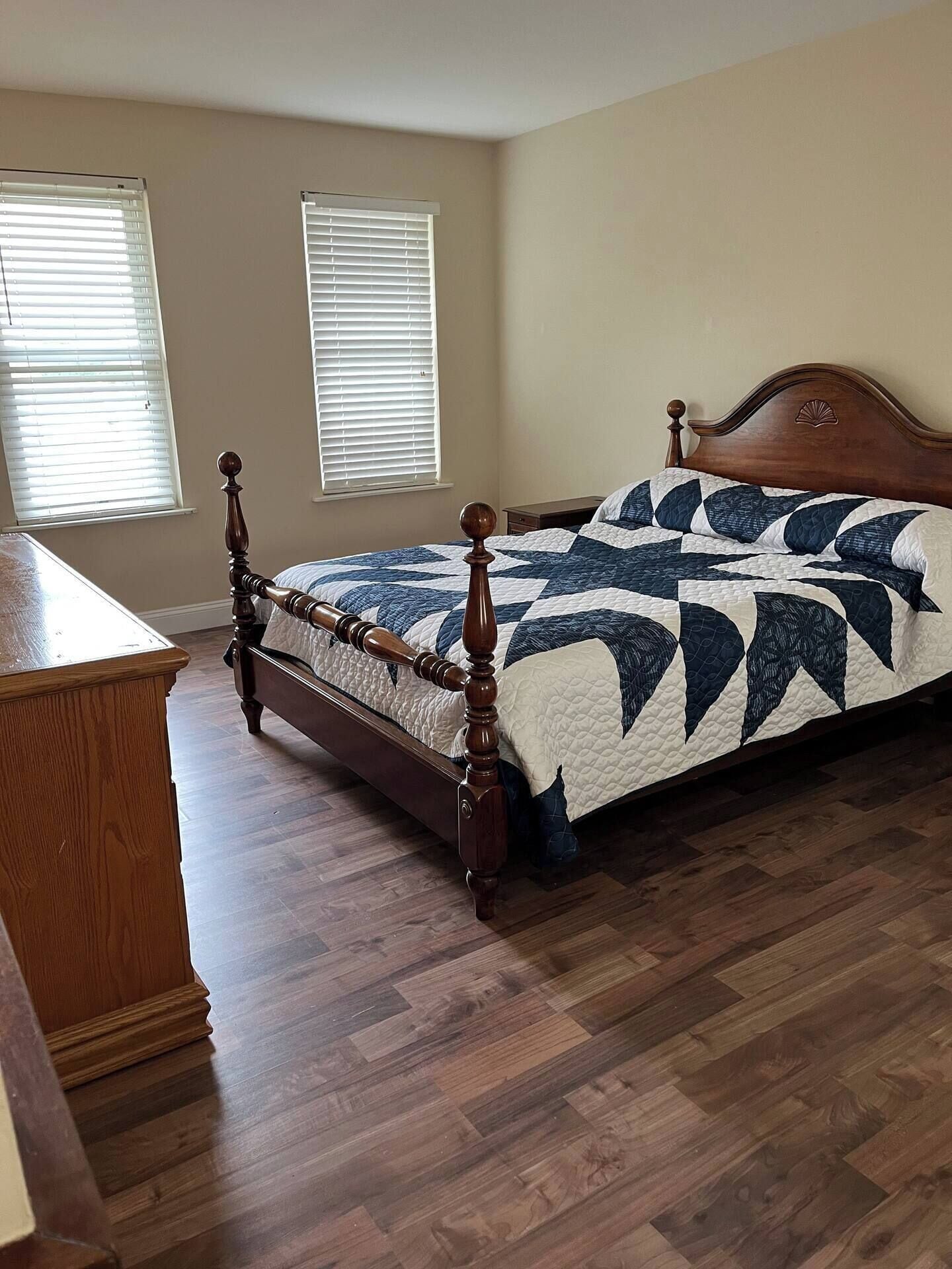 A bedroom with a wooden bed, a dresser, and two windows with blinds.