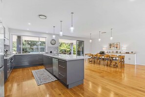 A modern kitchen with grey cabinets, a large island, and a dining area with wooden chairs. - Hamptons Estate: Freshwater Pool, 8 Mins Beach (Terrigal)