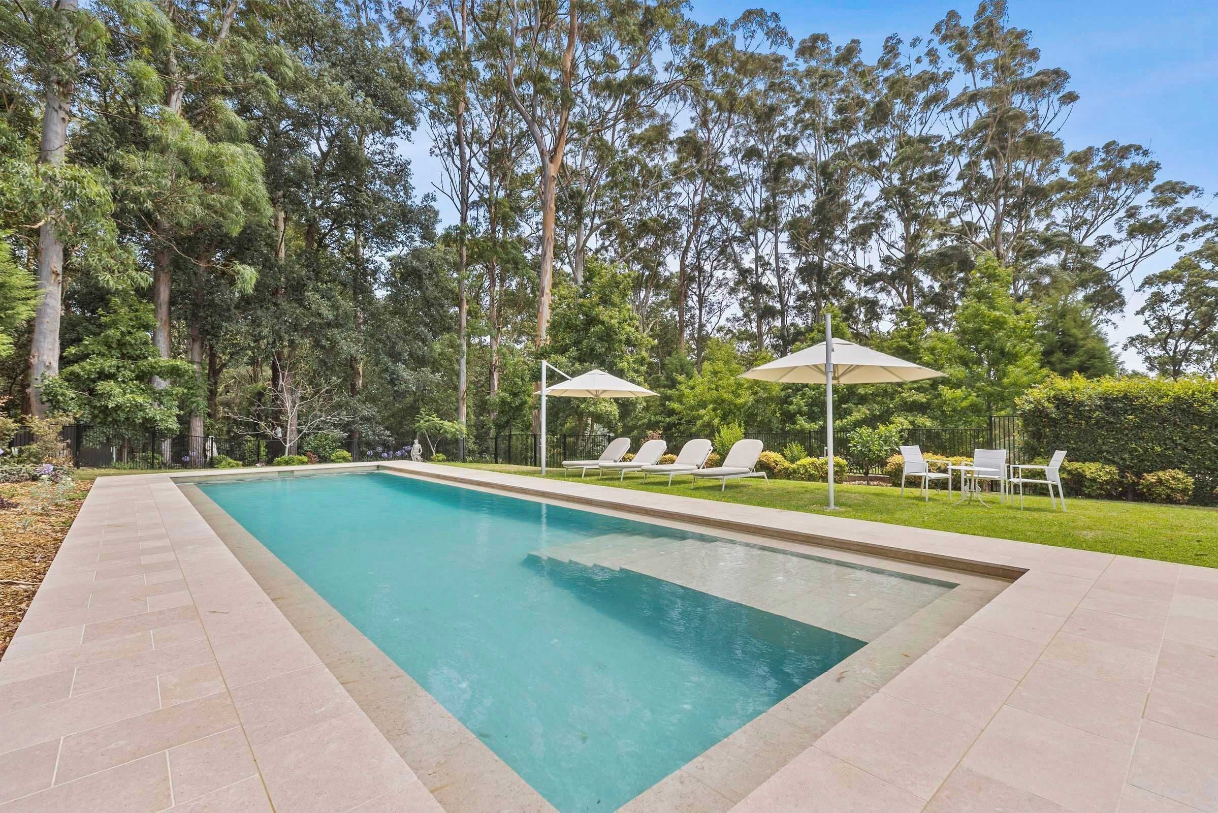 A rectangular swimming pool with a tiled edge, surrounded by a grassy area and trees. There are lounge chairs and umbrellas along the poolside.