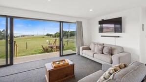 A modern living room with a large sliding glass door leading to a grassy outdoor area, a grey sofa, a wooden coffee table, and a flat-screen TV mounted on the wall.