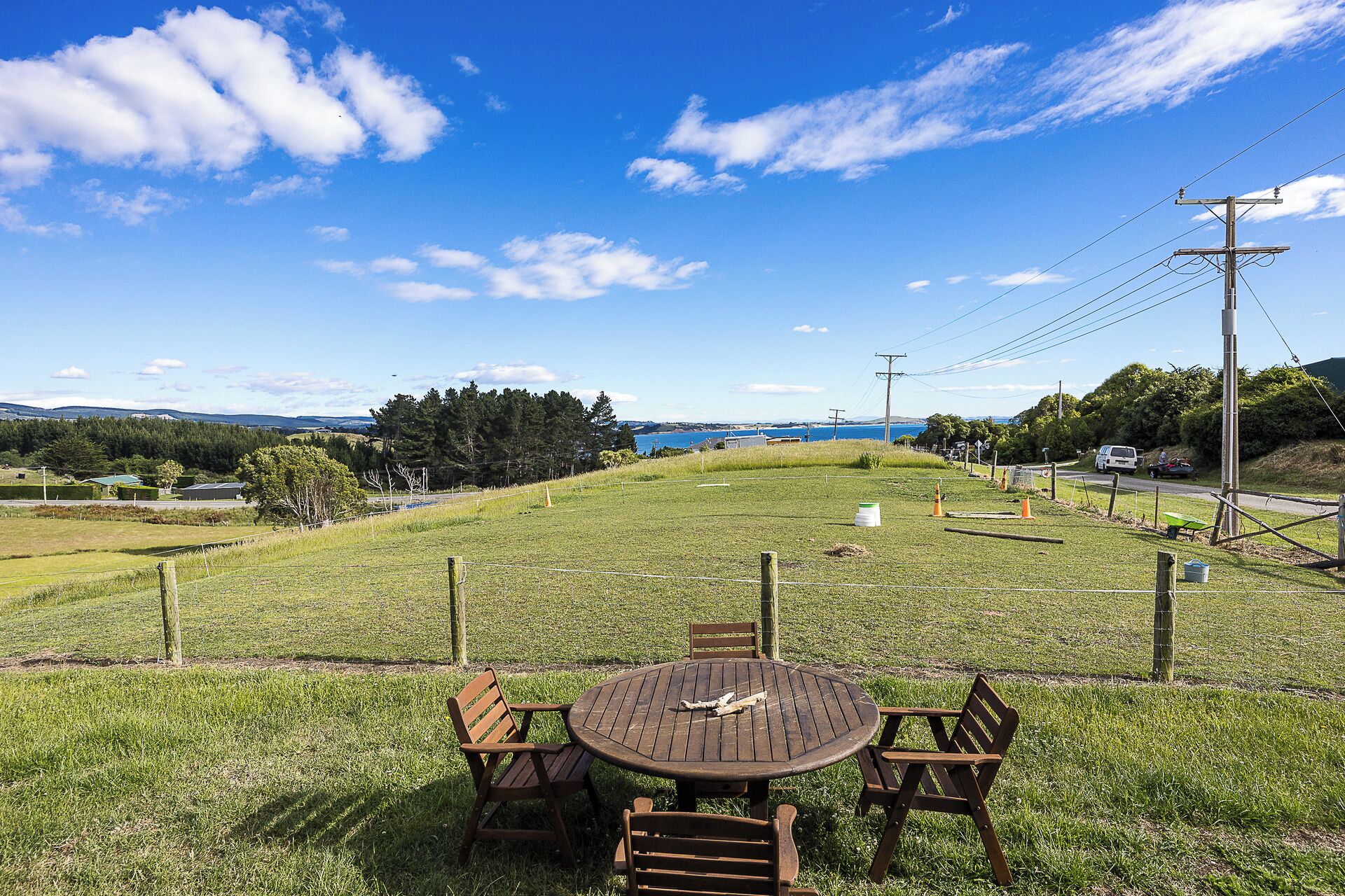 Un espace extérieur verdoyant avec une table et des chaises en bois, un terrain clôturé et une vue sur une étendue d’eau et des collines lointaines.