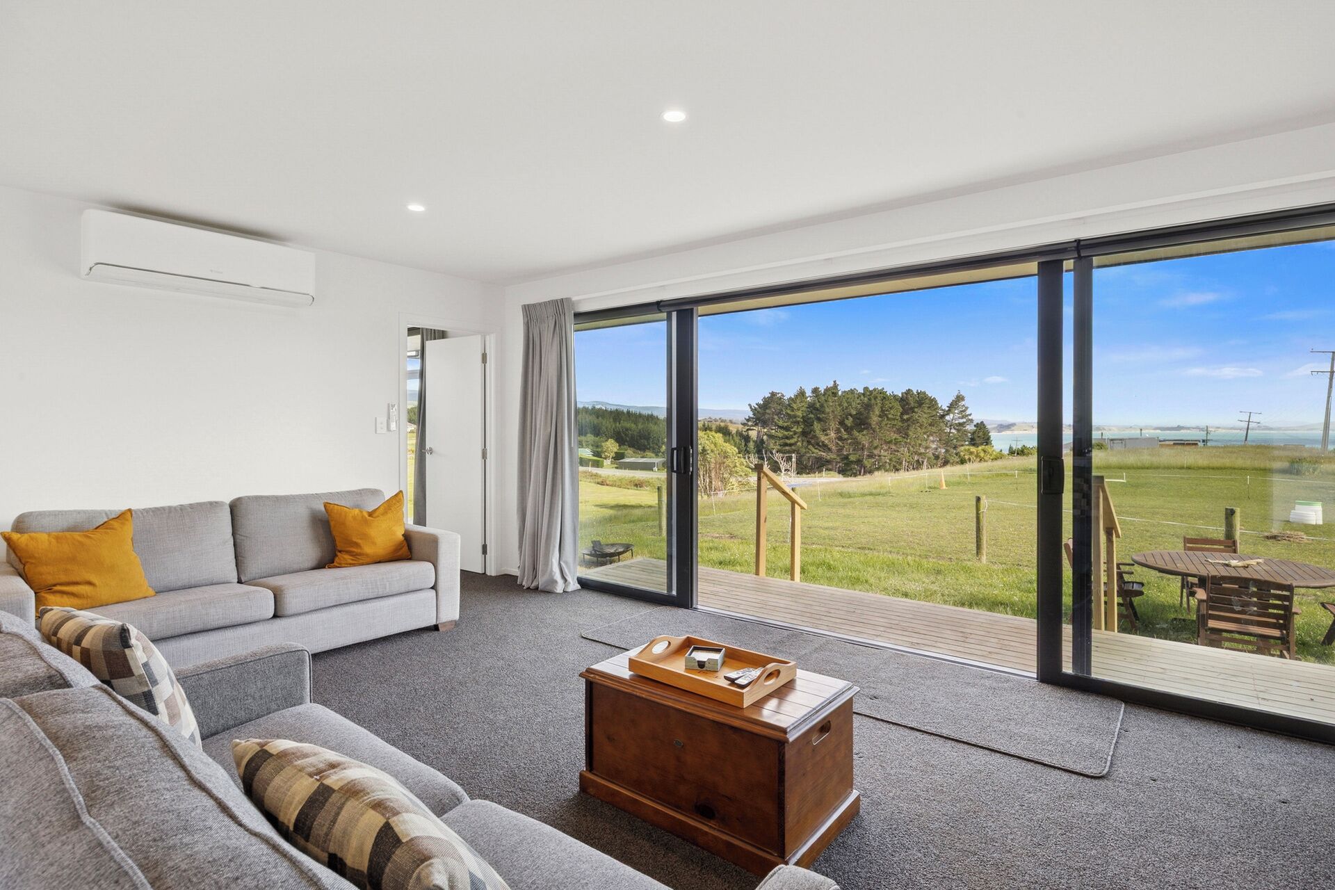 A modern living room with a grey sofa, a wooden coffee table, and a large sliding glass door leading to a deck with a view of a grassy landscape and a body of water.