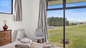 A bedroom with a large window, a bed with folded towels, two chairs, a wooden dresser, and a vase with dried flowers.