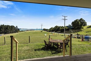 Eine grüne Außenanlage mit Holztisch und -stühlen, einem umzäunten Bereich und Blick auf Meer und Hügel im Hintergrund.
