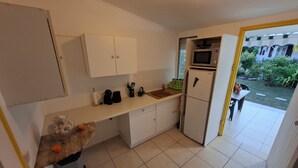 Cocina compacta con armarios blancos, un microondas y un refrigerador. Se ve un lavamanos pequeño y una encimera, con vista a un jardín exterior.