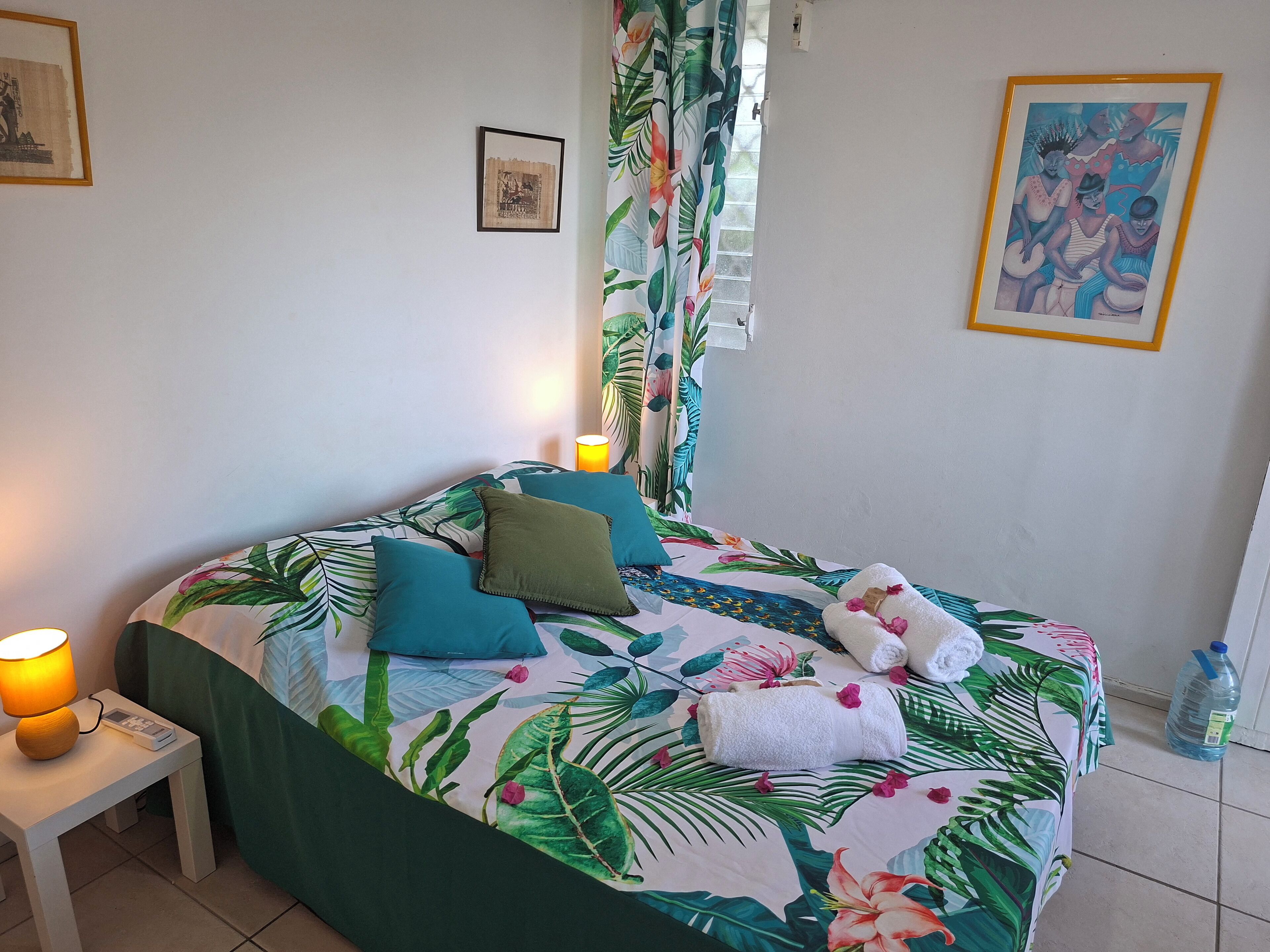 Un lit orné d’un motif de feuilles tropicales, des oreillers, une table de chevet avec une lampe et un tableau encadré accroché au mur.