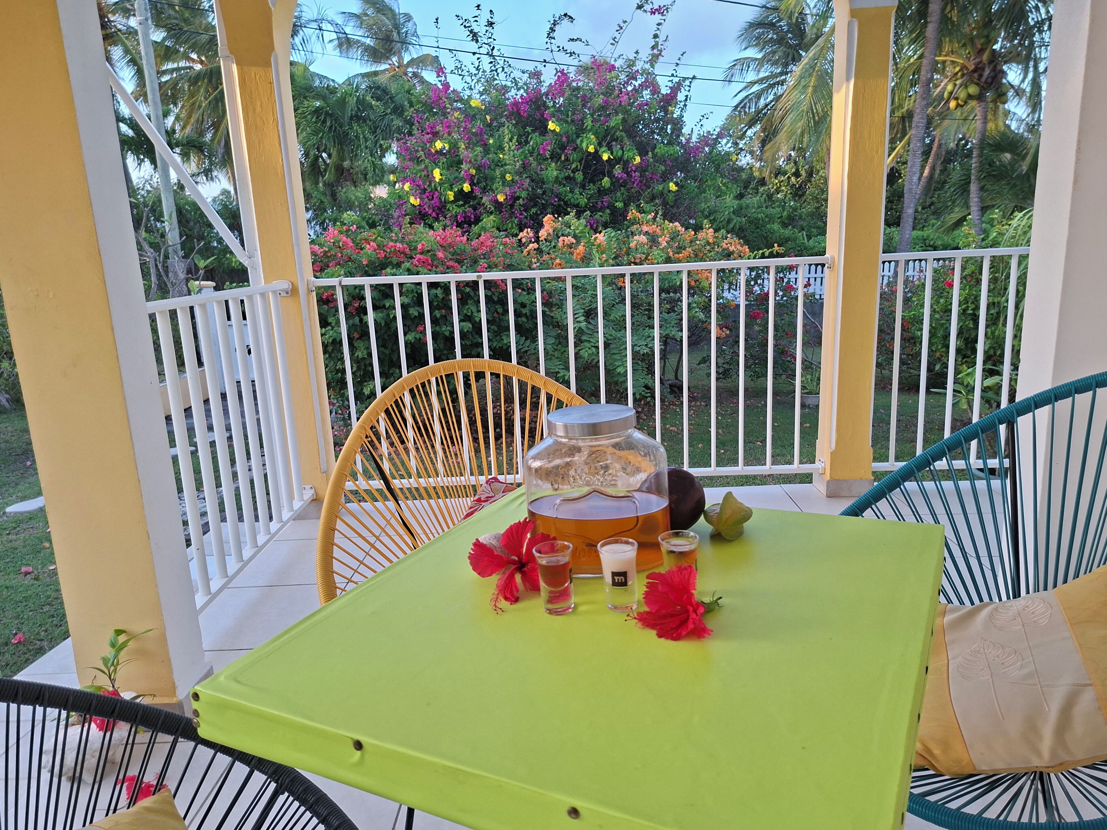 A balcony with a yellow table, a wicker chair, and a view of a garden with colorful flowers and palm trees.