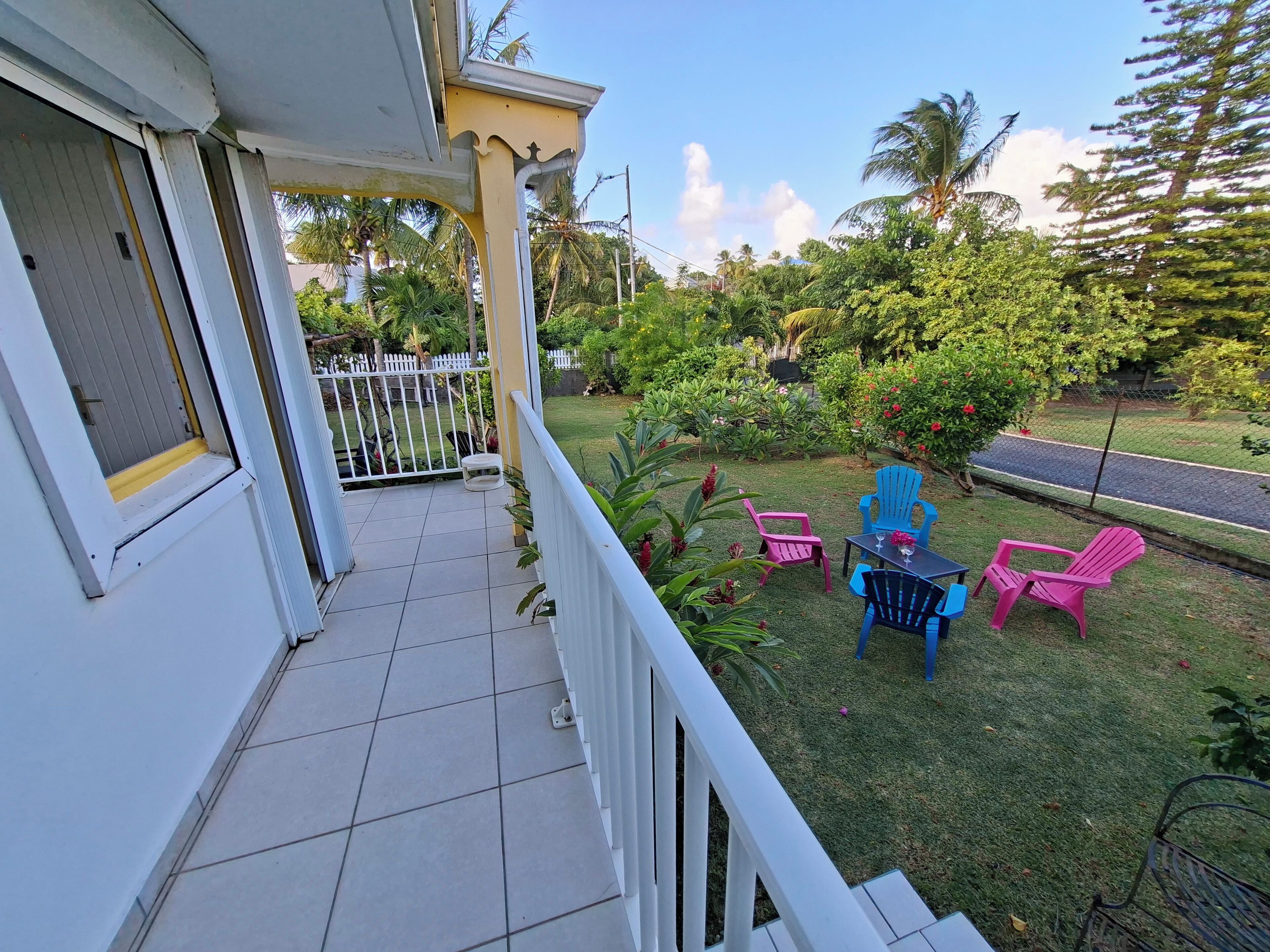 A balcony with a white railing, a small table with chairs, and a view of a lush garden with various plants and a white fence.