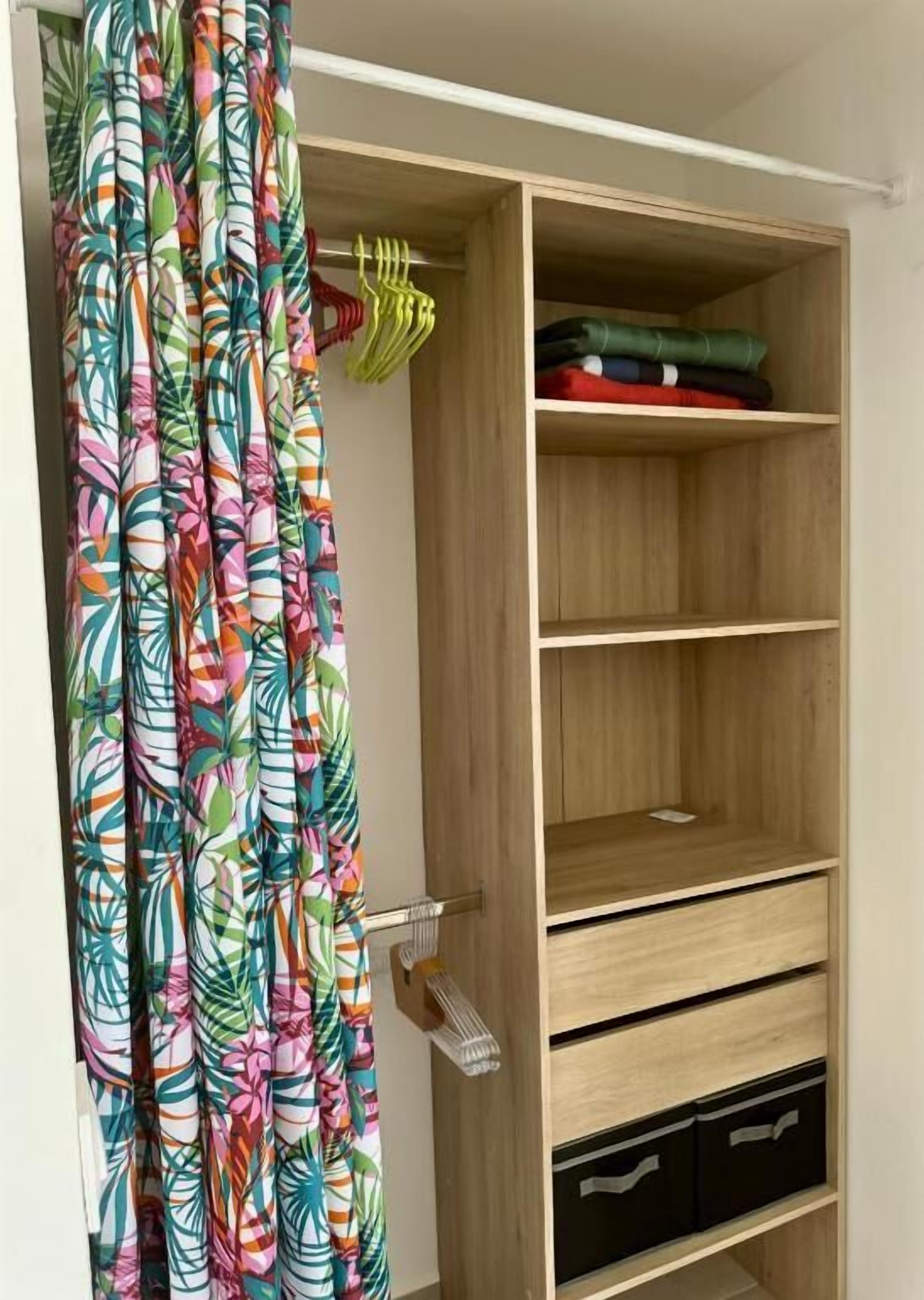 A wooden wardrobe with multiple shelves and drawers, a hanging rack with clothes, and a curtain with a tropical leaf pattern.