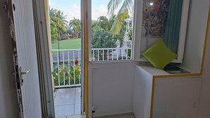 Una habitación con balcón, un ventanal pequeño con un cojín verde y vistas a un jardín con palmeras.