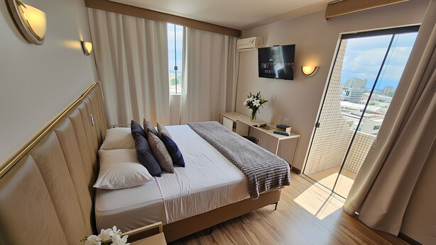 A modern hotel room with a large bed, a desk, a TV, and a balcony with a city view. - Santoni Hotel Guarapuava (Guarapuava)
