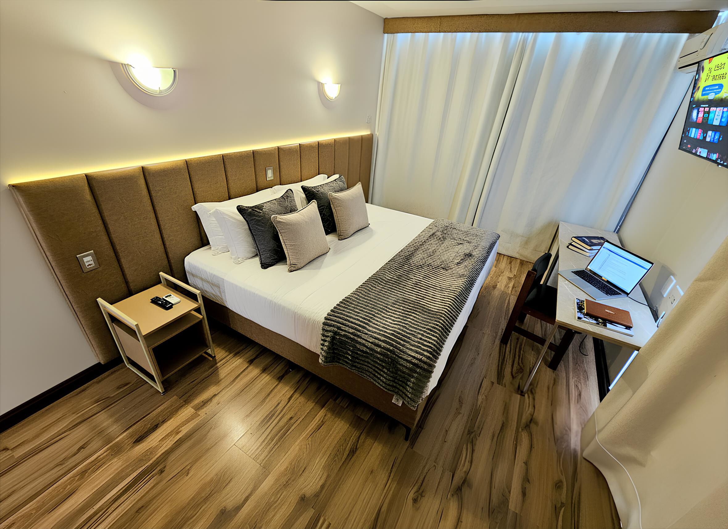 A modern hotel room with a large bed, bedside tables, a desk with a laptop, and a large window with curtains.
