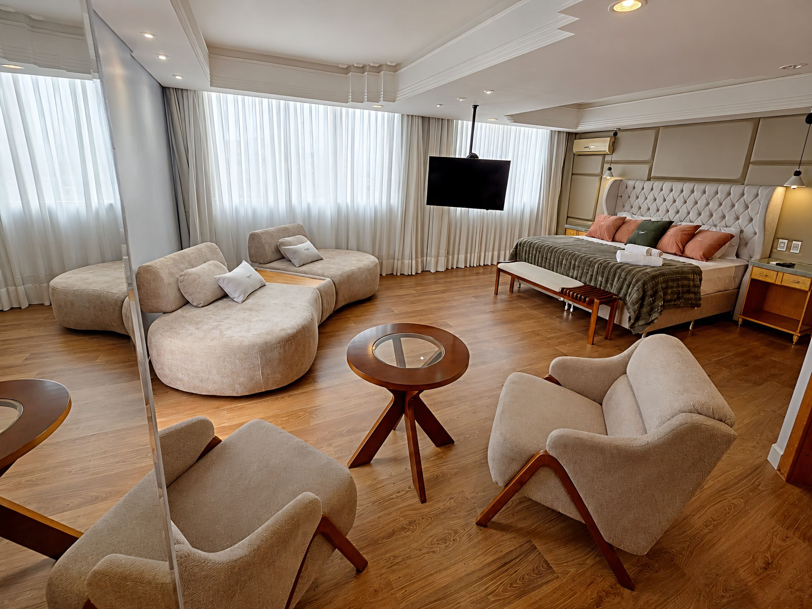 Una habitación de hotel moderna con un sofá curvo, una mesa de centro redonda, una cama con cabecera capitonada y un televisor de pantalla plana montado en la pared.