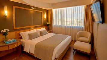 Quarto de hotel com uma cama grande, uma mesinha de cabeceira com um vaso de flores, uma televisão de tela plana na parede e uma janela com cortinas.