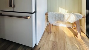 A modern kitchen with a white refrigerator, wooden flooring, and a small wooden stool with a white furry seat.
