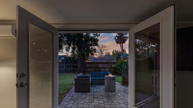 An open sliding glass door leading to a patio with a fire pit, outdoor furniture, and a view of a backyard with a fence and trees. - Happy Ours (New Smyrna Beach)