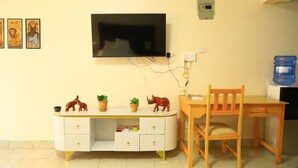 Ein Zimmer mit an der Wand befestigtem Fernseher, einem Holztisch mit Stuhl, einem Wasserspender und einer Kommode mit Schubladen und Dekoration.