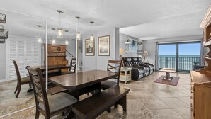 A spacious living area with a dining table, chairs, and a sofa. There is a view of the ocean through large windows. - Zodiac 4 (Daytona Beach Shores)