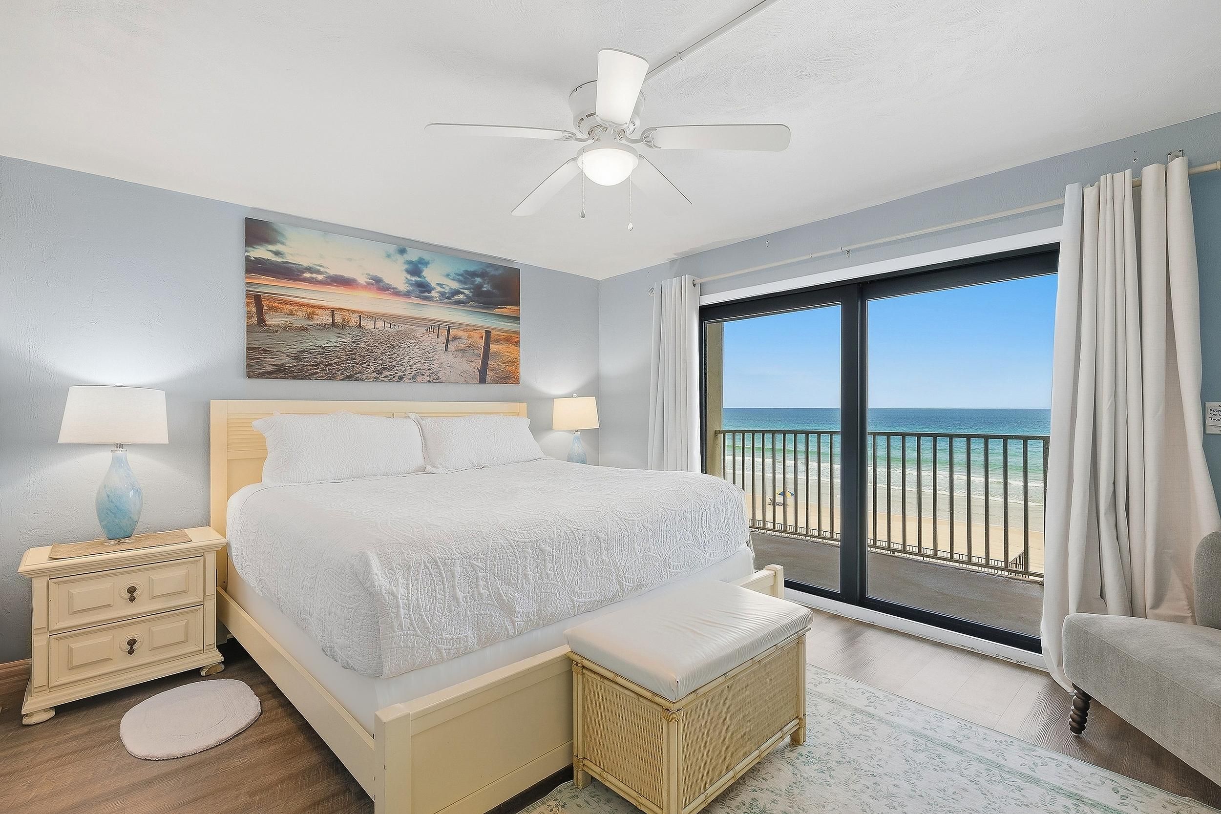 A bedroom with a bed, a nightstand, a lamp, a painting, and a view of the beach.