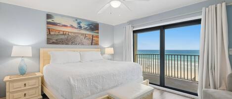 A bedroom with a bed, a nightstand, a lamp, a painting, and a view of the beach.