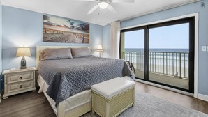A bedroom with a bed, nightstand, lamp, and a view of the beach through a large window. - Zodiac 4 (Daytona Beach Shores)