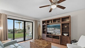 Ein modernes Wohnzimmer mit einem großen Flachbildfernseher, einem Deckenventilator und Blick ins Freie durch große Schiebeglasüren.
