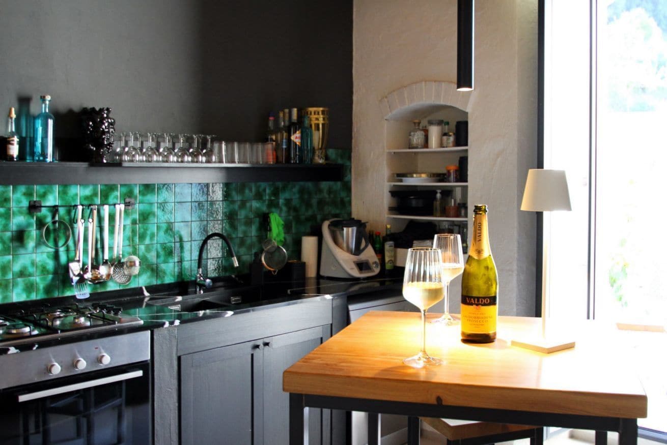 Una cucina con zoccolo verde piastrellato, un tavolo di legno con una bottiglia di vino e un bicchiere, e diversi utensili da cucina.