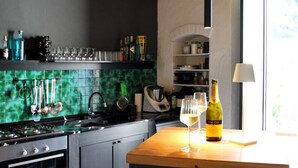 Ett kök med gröna kakelplattor på väggen bakom arbetsbänken, ett träbord med en vinflaska och ett vinglas samt diverse köksredskap.