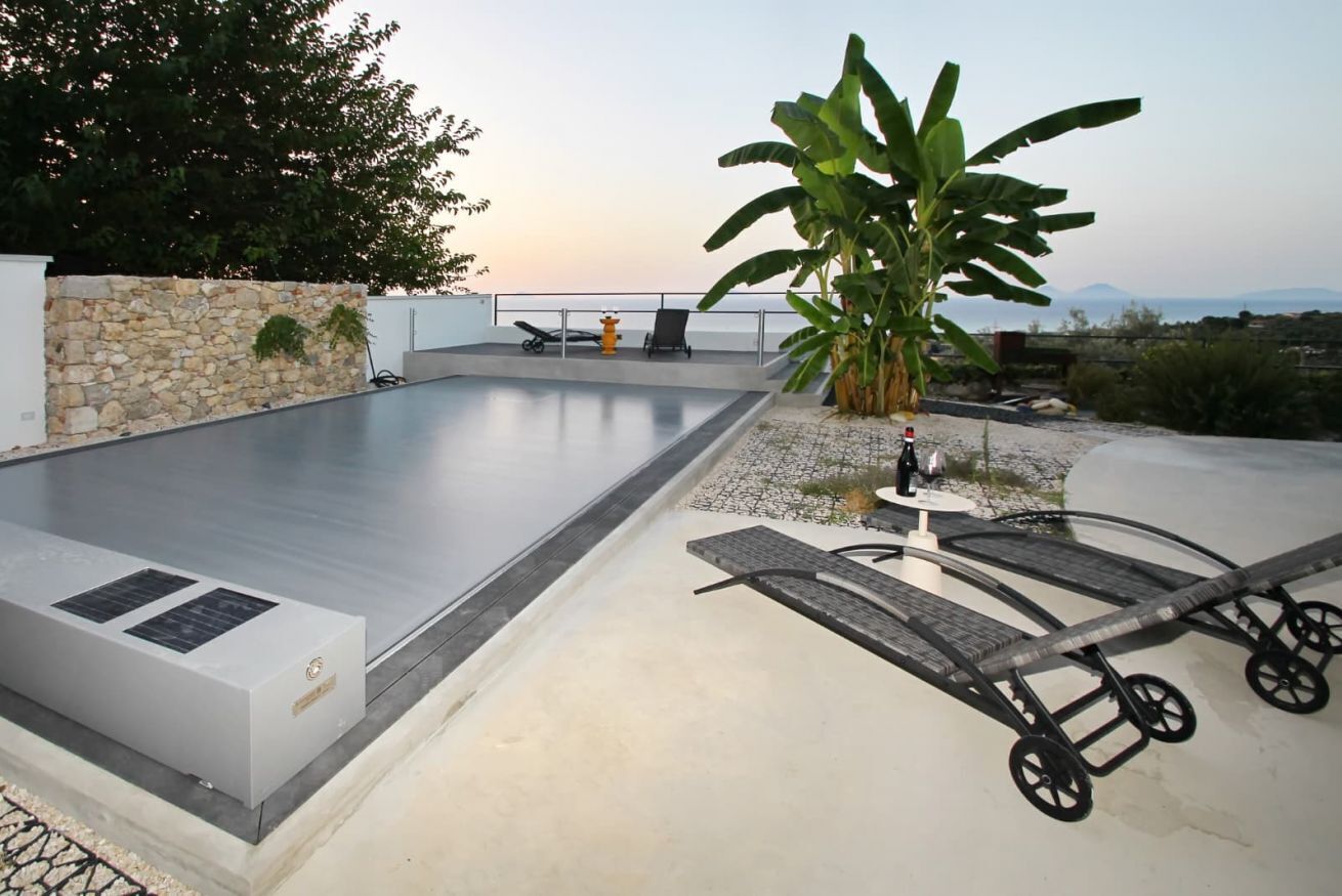A swimming pool with a stone wall and a large tree, surrounded by lounge chairs and a table.