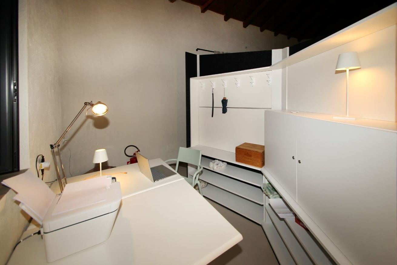 Una stanza compatta e moderna con una scrivania bianca, un portatile, una lampada e una sedia. È presente un armadio bianco con organizer pensili e un piccolo ripiano.