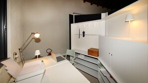 Ett kompakt och modernt rum med ett vitt skrivbord, en bärbar dator, en lampa och en stol. Det finns en vit garderob med en hängorganisatör och en liten hylla.